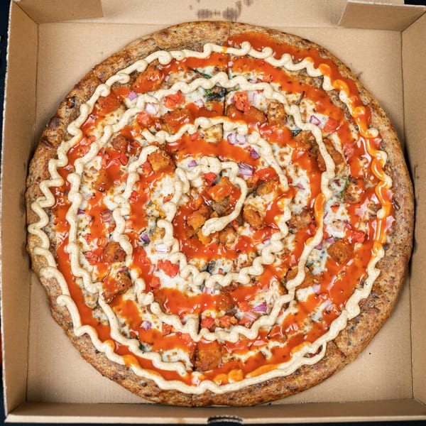 A vegan pizza with buffalo sauce, ranch drizzle, plant-based chicken, red onions, and tomatoes in a box.