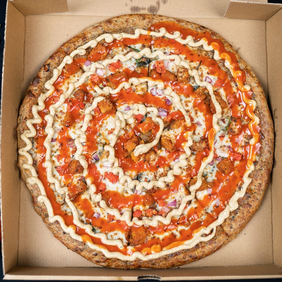 A vegan pizza with buffalo sauce, ranch drizzle, plant-based chicken, red onions, and tomatoes in a box.