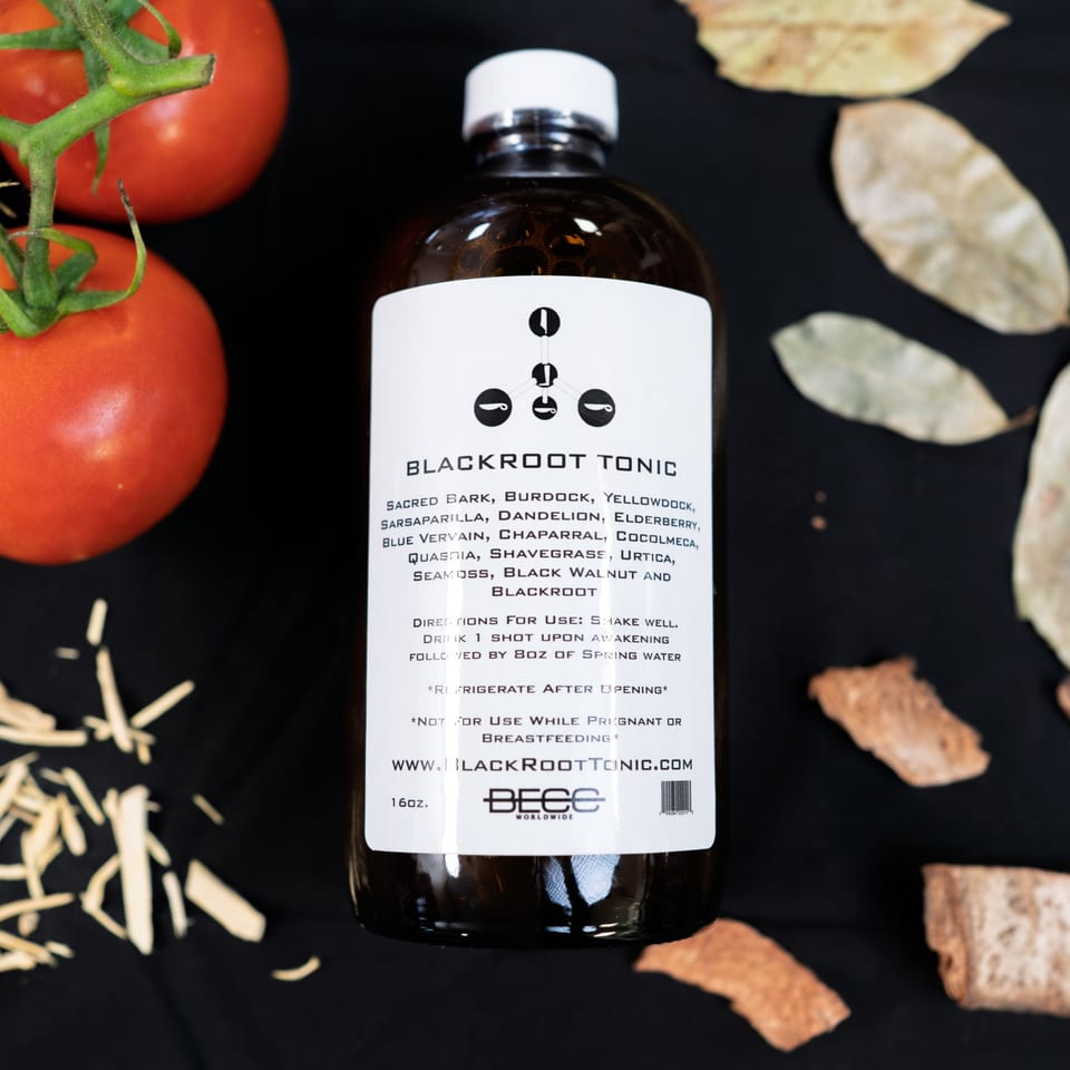 A bottle of Blackroot Tonic with tomatoes, wood shavings, and dried leaves on a black surface.