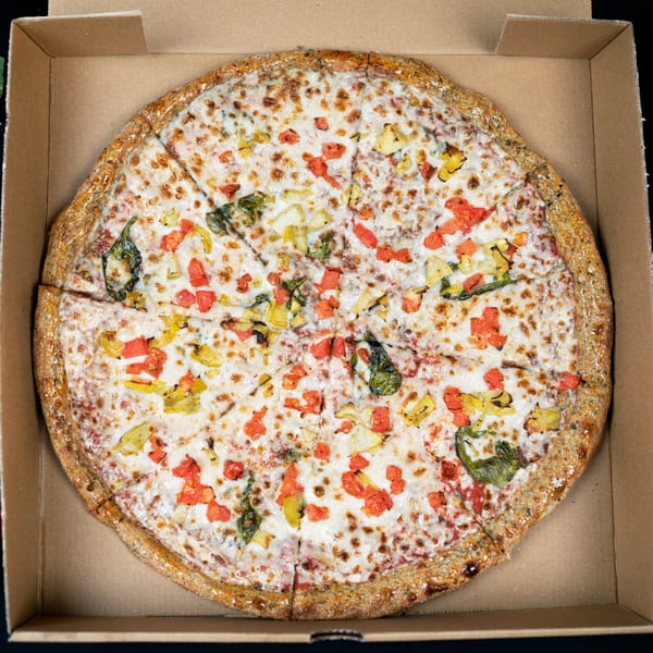 Whole vegan pizza with white cheese, red and yellow peppers, and basil in a cardboard box.