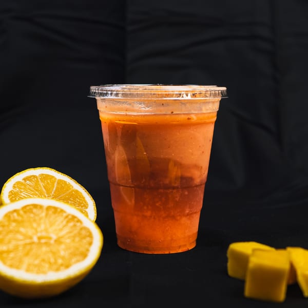 A vegan mango smoothie in a plastic cup with lemon halves and mango chunks on a black background.