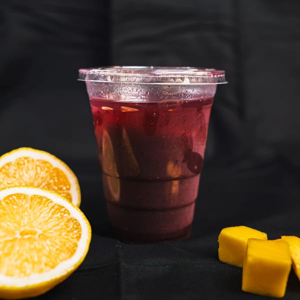 A vibrant red vegan smoothie in a clear cup, with orange slices and mango cubes on a dark surface.
