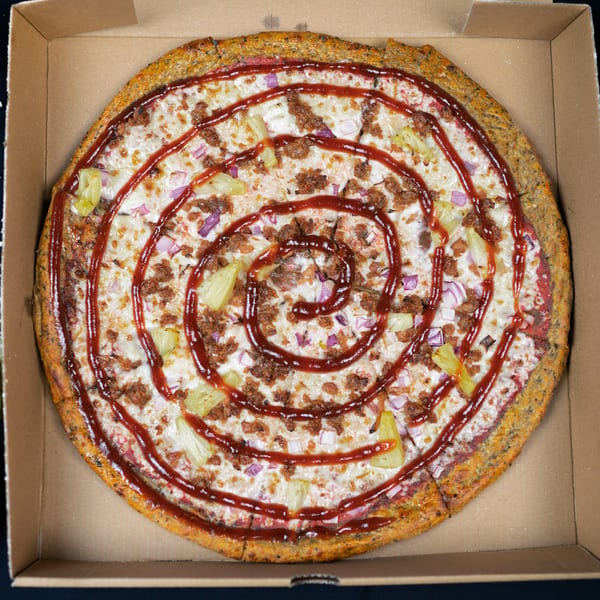 Vegan pizza with white sauce, plant-based meat, pineapple, red onion, and BBQ sauce swirl in a box.